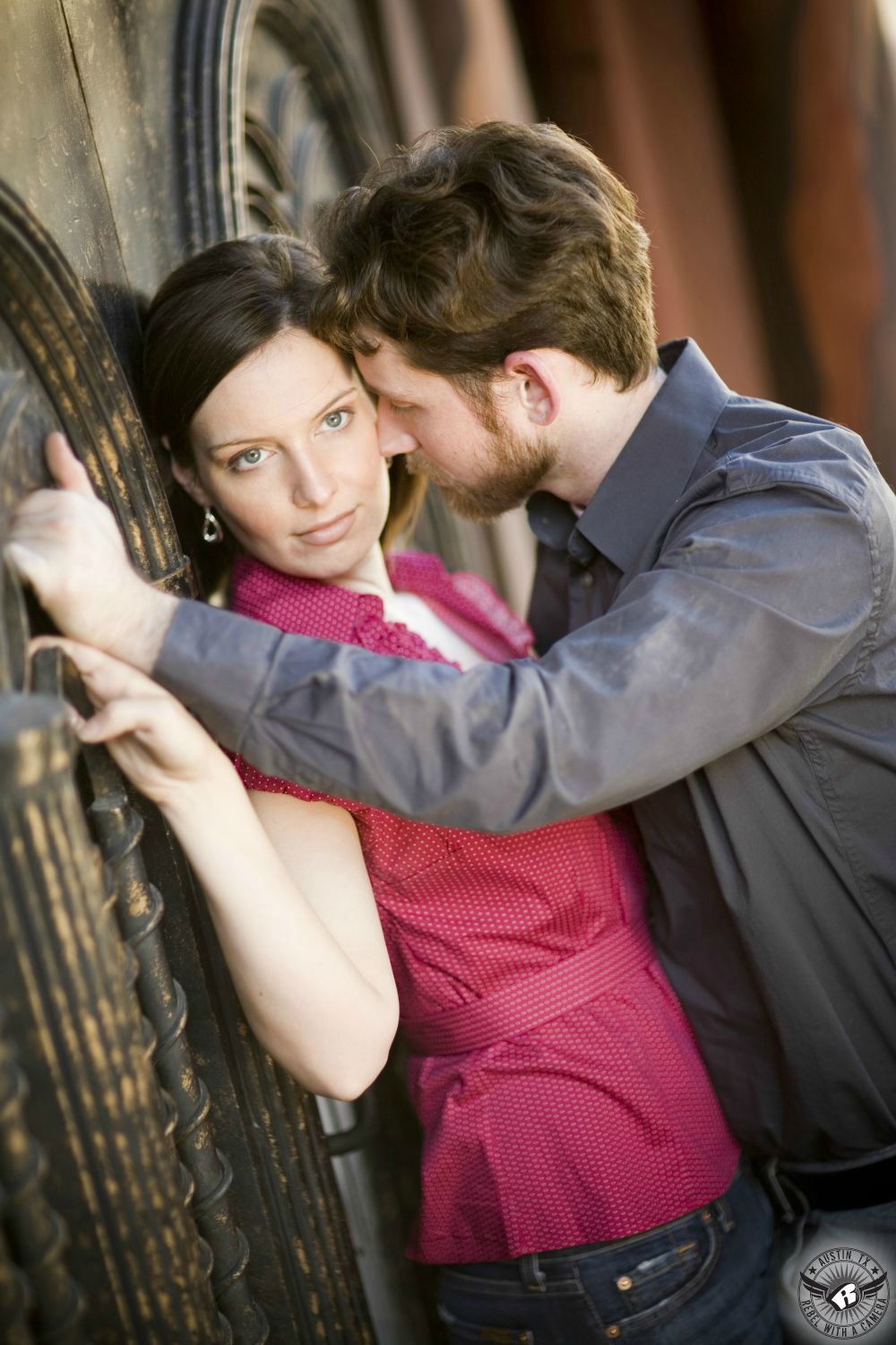 Blue eyed, brown hair girl wearing a pink low cut blouse with blue jeans is pressed against a gothic styled door by a brunette bearded guy wearing a grey long sleeve button up shirt in this intense engagement portrait in the warehouse district of Austin.